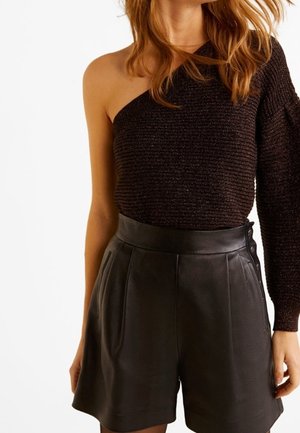 Vrouw draagt een zwart topje met één schouder en lange mouwen, gecombineerd met zwarte leren shorts met hoge taille, tegen een witte achtergrond.