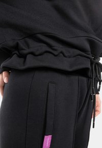 Completo sportivo nero con elastico in vita. Presenta un tessuto testurizzato, chiusure con coulisse e un piccolo etichetta logo viola sul lato.