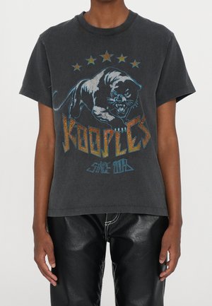 Persoon draagt een vervaald zwart T-shirt met een grafiek van een grommende panter, vijf sterren erboven en de tekst "Kooples Since 2008" op de voorkant.