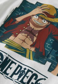 Balta marškinėliai su spalvinga grafika, vaizduojančia animacinio personažo, dėvinčio šiaudinę skrybėlę ir raudoną švarką, šviesias spalvas ir detalius raštus.