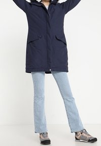 Marinblå parkas med foder av päls, dragkedja fram, två sidofickor och justerbara ärmslut. Bärs med ljusblå utsvängda jeans och grå skor.