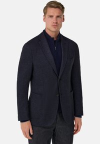 Tmavě modrý texturovaný blejzr s jedním knoflíkem, výřezovými klopami, kapsami s klopou a kontrastním tmavým zipem, v kombinaci s tmavými kalhotami.