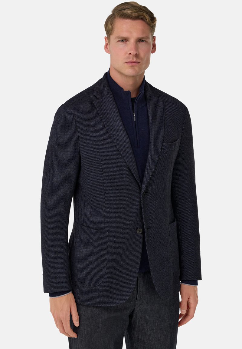 Tmavě modrý texturovaný blejzr s jedním knoflíkem, výřezovými klopami, kapsami s klopou a kontrastním tmavým zipem, v kombinaci s tmavými kalhotami.