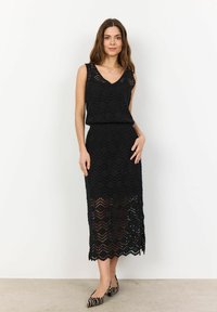 Abito in crochet nero con scollatura a V, senza maniche, caratterizzato da un motivo a zigzag e spacco laterale. Abbinato a ballerine a righe bianche e nere.