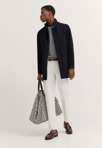 Marineblaue wasserdichte Jacke, gestreiftes weißes Hemd, weiße Hose, braune Slipper, trägt eine gemusterte Tragetasche mit geometrischem Design.