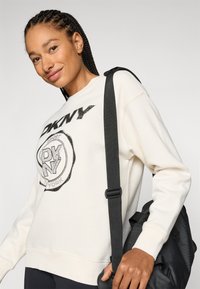 Felpa color crema con logo e grafica nera DKNY, dotata di polsini e orlo a costine. La tracolla nera di una borsa è visibile su uno sfondo semplice.