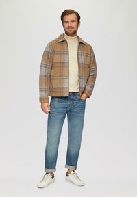 Jacket w kratę w odcieniach brązu i szarości, beżowy golf, niebieskie dżinsy oraz białe trampki. Kurtka ma kołnierz i dwie przednie kieszenie.