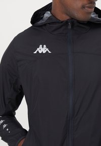 Sort letvægtsjakke med lynlås foran, hætte og Kappa-logo i hvid på brystet. Har elastiske manchetter og en glat stofoverflade.