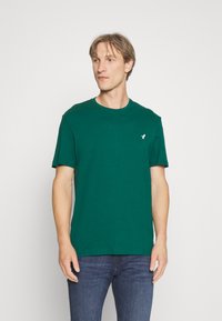 Dunkelgrünes Baumwoll-T-Shirt mit Rundhalsausschnitt und kurzen Ärmeln. Mit einem kleinen weißen Logo auf der linken Brustseite. Lässige Passform.