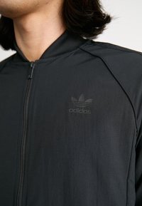 Juodas užtrauktukas striukė su lygiu paviršiumi ir prigludusiu dizainu, turinti subtilų juodą Adidas logotipą kairėje krūtinės pusėje.