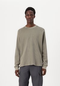 Olivengrünes Langarmshirt aus strukturiertem Stoff, mit Rundhalsausschnitt und Raglanärmeln. Kombiniert mit grauen Cargo-Hosen.