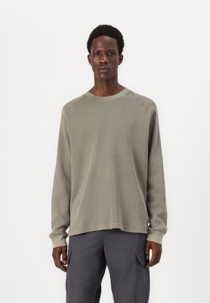 Olive green long-sleeve shirt in textured fabric, featuring a crew neckline and raglan sleeves. Paired with gray cargo pants.