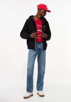 Uomo con cappellino rosso, t-shirt rossa con grafica, felpa nera con zip, jeans blu e scarpe da ginnastica bianche, in piedi davanti a uno sfondo bianco semplice.