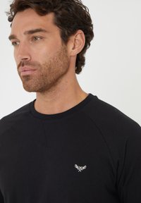 Sweatshirt noir avec un col rond et des manches raglan. Il présente un petit logo blanc brodé sur la poitrine. Tissu en coton lisse.