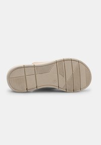 Semelle de chaussure avec une surface en caoutchouc beige texturée, dotée de rainures courbées et droites pour une meilleure adhérence. Logo circulaire au niveau du talon.