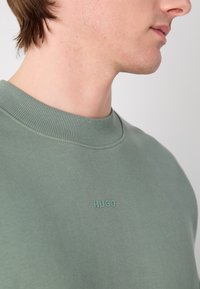 Sudadera de algodón verde con cuello redondo, con un sutil logo en relieve "HUGO" en el pecho. Textura suave y diseño minimalista.