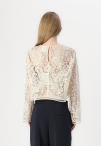 Haut en dentelle à manches longues, couleur ivoire, présentant un dos en découpe en cœur et un ourlet festonné, superposé sur un pantalon sombre.