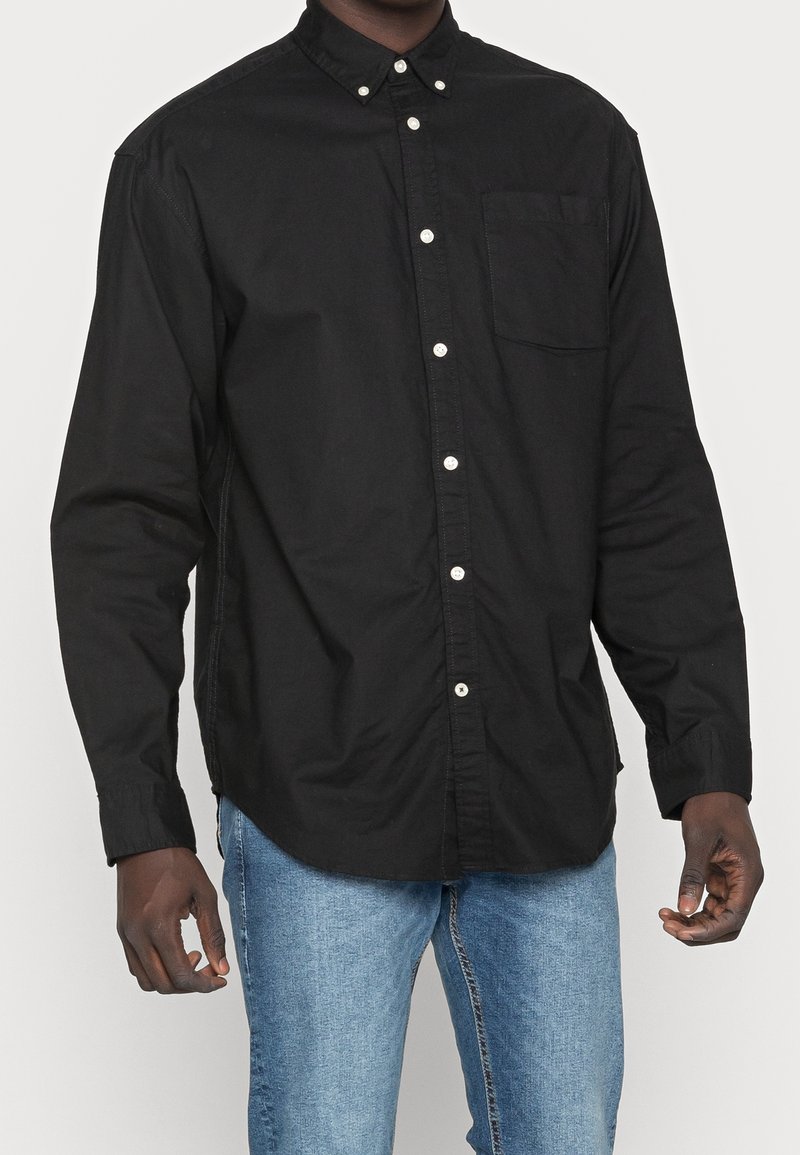 Camicia nera con bottoni realizzata in cotone. Caratterizzata da colletto, maniche lunghe e taschino sul petto. I bottoni sono bianchi, creando un contrasto con il tessuto.