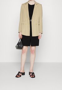 Woman wearing beige blazer over black dress with front slit, holding black handbag, wearing black heeled sandals with braided straps.