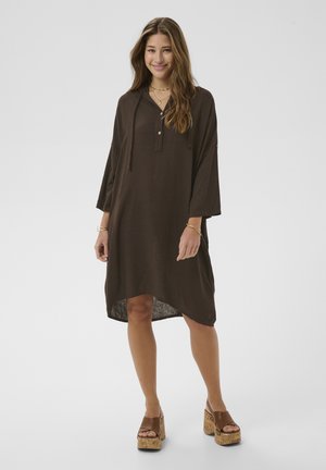 Mladá žena sa usmieva, nosí voľné hnedé šaty s rukávmi a hnedé sandále na platforme, stojí na jednoduchom svetlom pozadí.