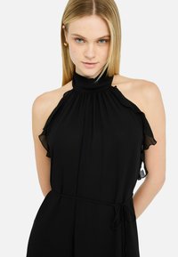 Crna halter haljina s volanima, izrađena od glatkog materijala. Ima naborani izrez i detalj u obliku zavezanog struka.