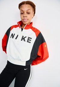 Balta, sarkana un melna Nike vēja jaka ar augstu apkakli, vienu pusi rāvējslēdzēju un pārspīlētu piegriezumu; apdare ar lielu melnu "NIKE" logotipu un elastīgu jostasvietu.