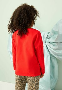 Rode lange mouwen shirt van katoen, gecombineerd met tan leopardenpatroon broek. Krullend haar en een lichtblauwe stofachtergrond.