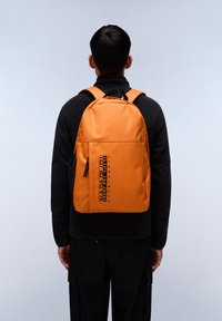 Zaino arancione con un logo nero, dotato di spallacci imbottiti e una tasca laterale con cerniera. Materiale liscio e forma rettangolare.