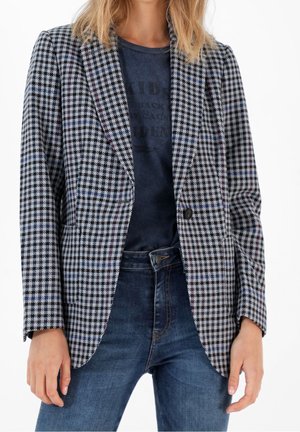 Femme portant un blazer pied-de-poule noir et blanc sur un t-shirt graphique foncé et un jean bleu, debout avec les mains détendues.