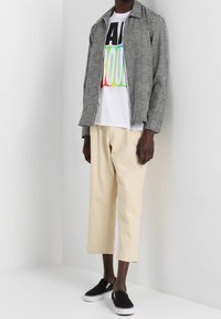 Veste à motif pied-de-poule noir et blanc, t-shirt blanc avec texte coloré, pantalon court beige clair et chaussures noires sans lacets.