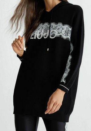 Femme portant un long sweat à capuche noir avec un motif en dentelle blanche et le texte « LIU JO », tenant un cordon près de son épaule.