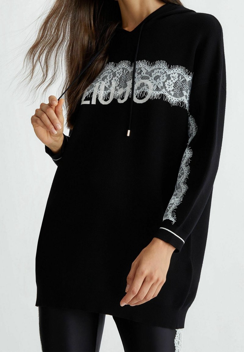 Femme portant un long sweat à capuche noir avec un motif en dentelle blanche et le texte « LIU JO », tenant un cordon près de son épaule.