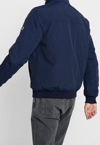 Mörkblå bomberjacka med quiltad textur, ribbade muddar och nederkant. Har en logotypapplikation på vänster ärm. Minimalistisk design.