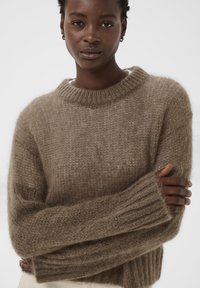 Pull oversize brun en laine avec un col rond, des poignets côtelés et une texture douce et duveteuse. Le mannequin pose avec les bras croisés.