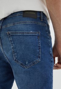 Pantalones vaqueros de mezclilla azul oscuro con un bolsillo trasero que muestra costuras visibles y una etiqueta negra en la cinturilla. La tela presenta una ligera textura.