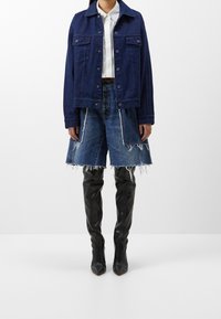 Laivastonsininen farkkutakki, jossa napit, yhdistettynä farkkuminihameeseen, jossa on repsottavat reunat, täydentäen asua mustilla reisiin ulottuvilla saappailla.