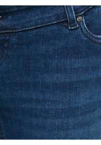 Close-up van donkerblauwe spijkerbroek met zicht op zakrand, ceintuurlus en metalen klinknagel op een gestructureerde stof.