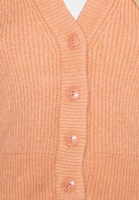 Persikofärgad stickad kofta med V-halsdesign, prydd med fem runda, genomskinliga knappar och ribbad textur genom hela plagget.