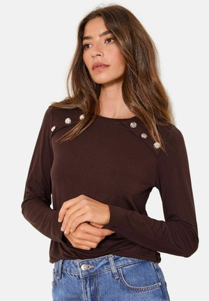 Woman with long brown hair wearing a dark brown long-sleeve top with silver buttons on shoulders and blue jeans, posing against white background.