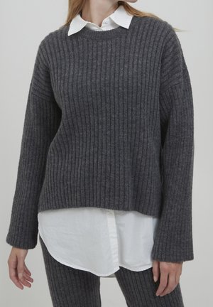 Persona che indossa un maglione a coste grigio scuro sovrapposto a una camicia bianca abbottonata e pantaloni a coste coordinati.