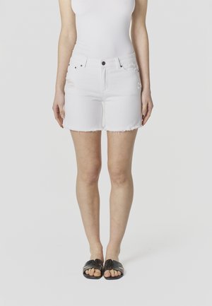 Vrouw met witte versleten denimshorts, wit nauwsluitend topje en zwarte slip-on sandalen, staand tegen een effen achtergrond.