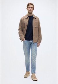 Φανελένιο πουκάμισο-ζακέτα σε ταμπά απόχρωση με δύο τσέπες στο στήθος, συνδυασμένο με μπλε σκούρο μπλουζάκι, ανοιχτόχρωμα τζιν και μπεζ αθλητικά παπούτσια. Μινιμαλιστικός σχεδιασμός.