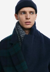 Wool coat with a dark navy and green plaid pattern, featuring a high collar and a soft, gray knitted scarf layered underneath.