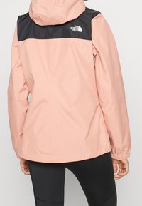 Veste imperméable rose clair, panneaux d'épaule noirs, poignets élastiques et capuche. Logo sur la partie supérieure du dos. Design épuré.