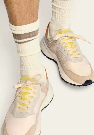 Des baskets claires, présentant un mélange de textures lisses et en daim, avec des lacets jaunes, des accents orange et une épaisse semelle noire. Associées à des chaussettes crème.