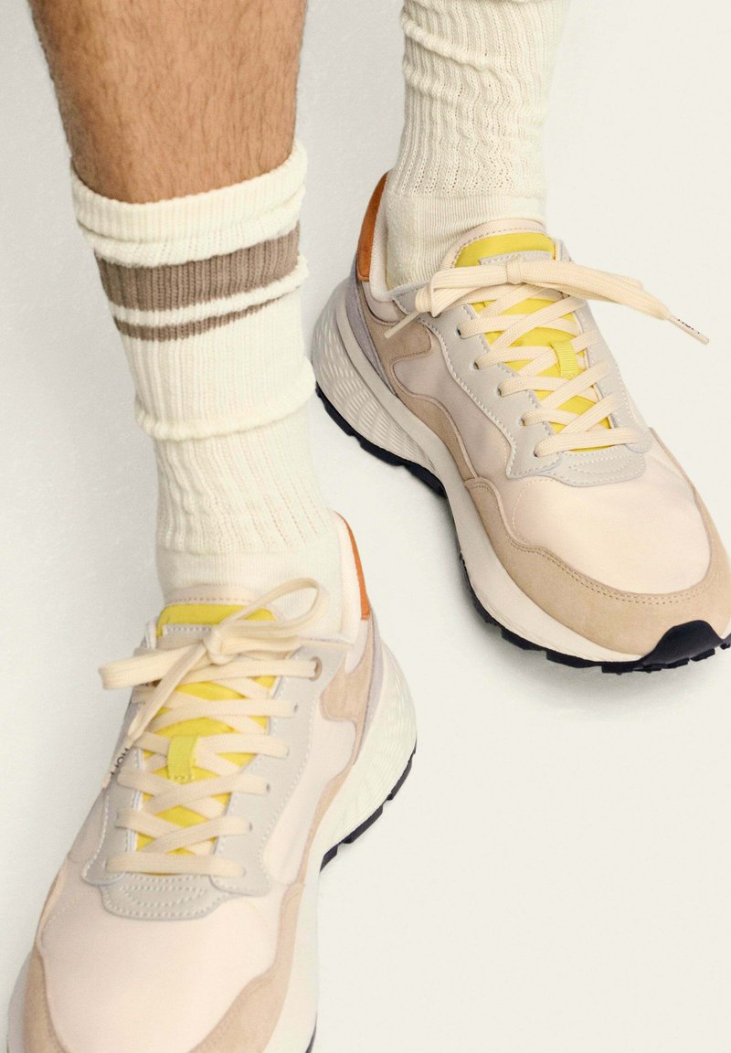 Des baskets claires, présentant un mélange de textures lisses et en daim, avec des lacets jaunes, des accents orange et une épaisse semelle noire. Associées à des chaussettes crème.