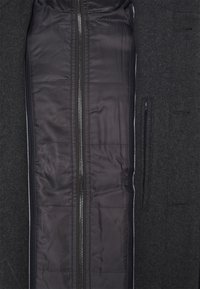 Gros plan d’un manteau gris foncé avec devant ouvert montrant la doublure intérieure noire matelassée et la fermeture éclair métallique visible.