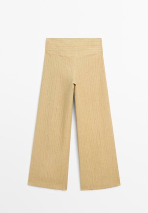 Beige bukser med vide ben med lodret ribbet tekstur og en høj, bred linning, vist fladt mod en hvid baggrund.