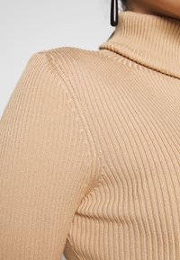 Maglione a collo alto color beige con coste, caratterizzato da un design aderente, una texture morbida e cuciture nette lungo le spalle e il colletto.