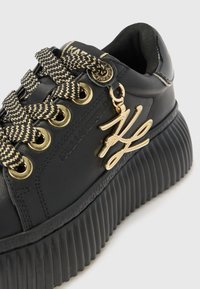 Sorte plattform sneakers med teksturert rille såle, gull aksenter, svarte og gull mønstrede snøringer, og en merket anheng festet.